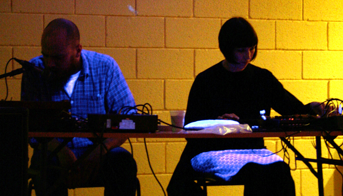 Dylan and Karen sit playing electronics silhouetted in front of a yellow wall