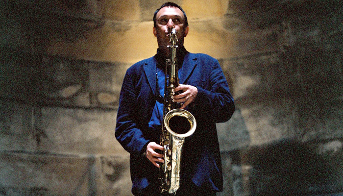 John butcher Plays saxophone direct to camera in front of a stone wall