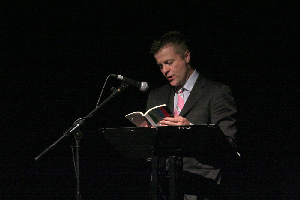 Christian Bok reads behind a lectern in a pink tie and suit