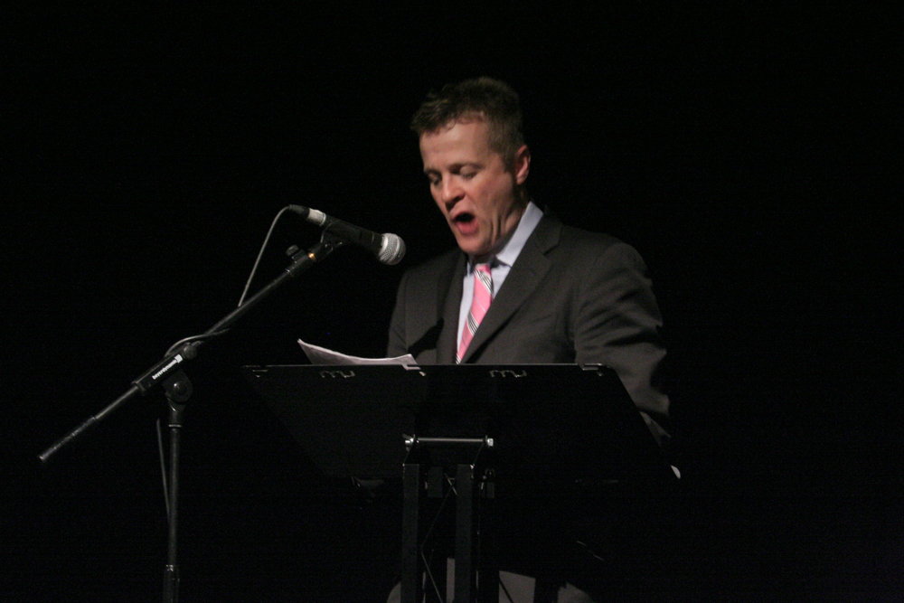 Christian Bok reads behind a lectern in a pink tie and grey suit