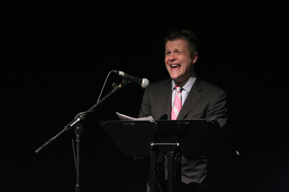 Christian Bok reads behind a lectern in a pink tie and grey suit