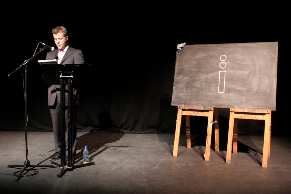 Christian Bok reads behind a lectern in a pink tie beside a blackboard
