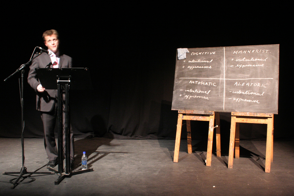 Christian Bok reads behind a lectern in a pink tie beside a blackboard