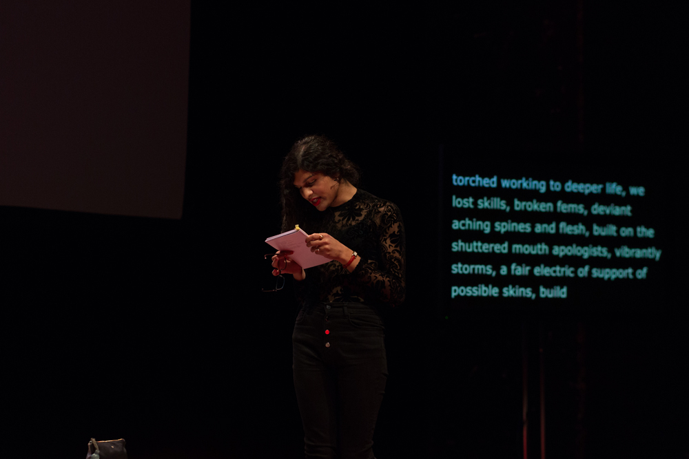 Nat Raha reading from a booklet on stage at EPISODE 9 
