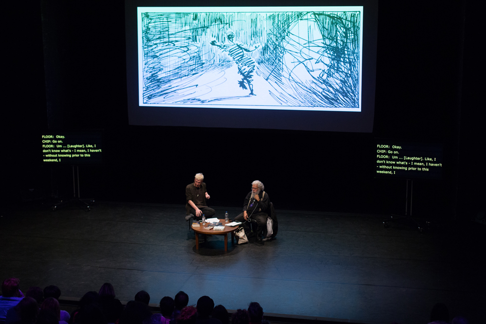 A screen showing a drawing behind Barry & Samuel on stage before an audience