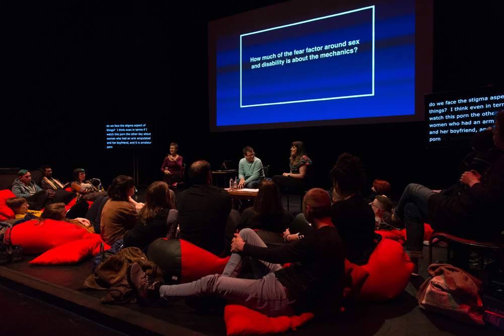 Two speakers on stage surrounded by audience and red cushions