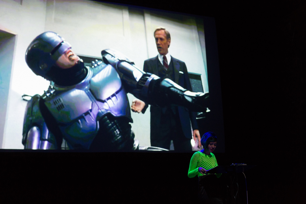 Robocop suffering on screen behind Jackie Wang reading on a stage