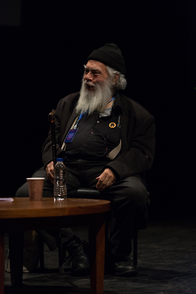 Bearded Samuel Delany wearing a little black hat smiling on stage by a table