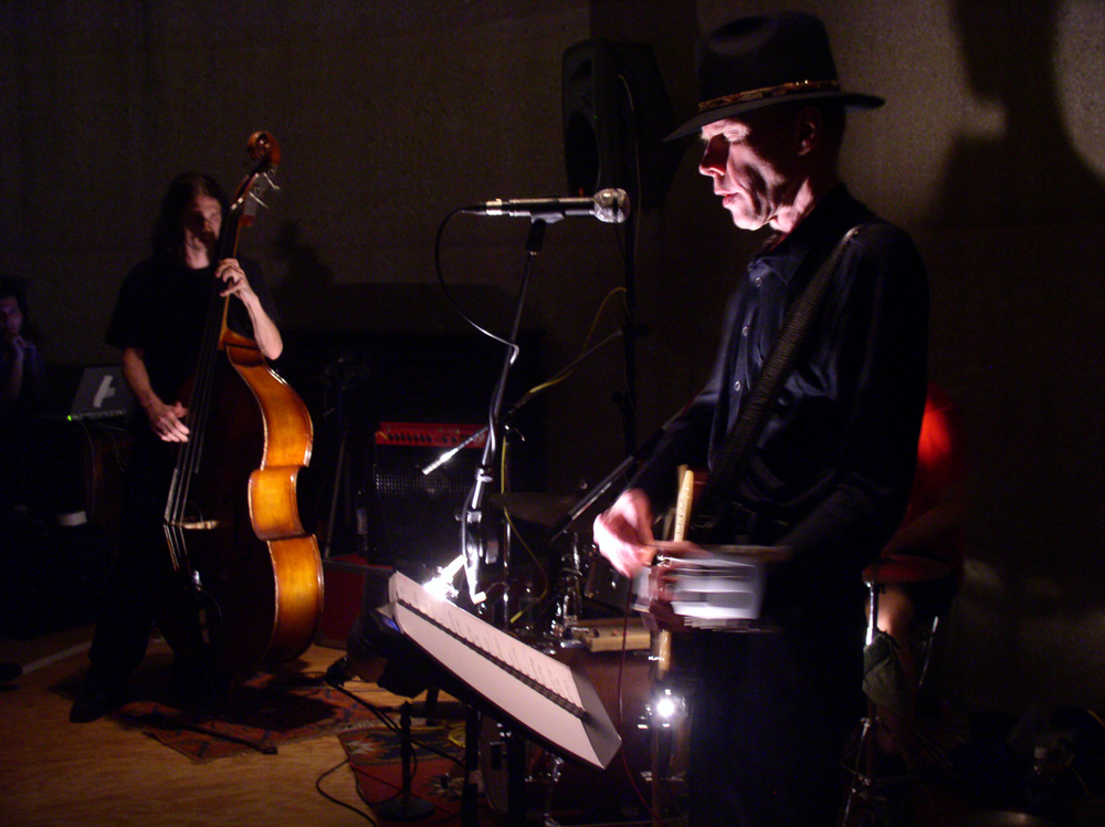 A skeletal Jandek playing a guitar with acoustic bass in the background