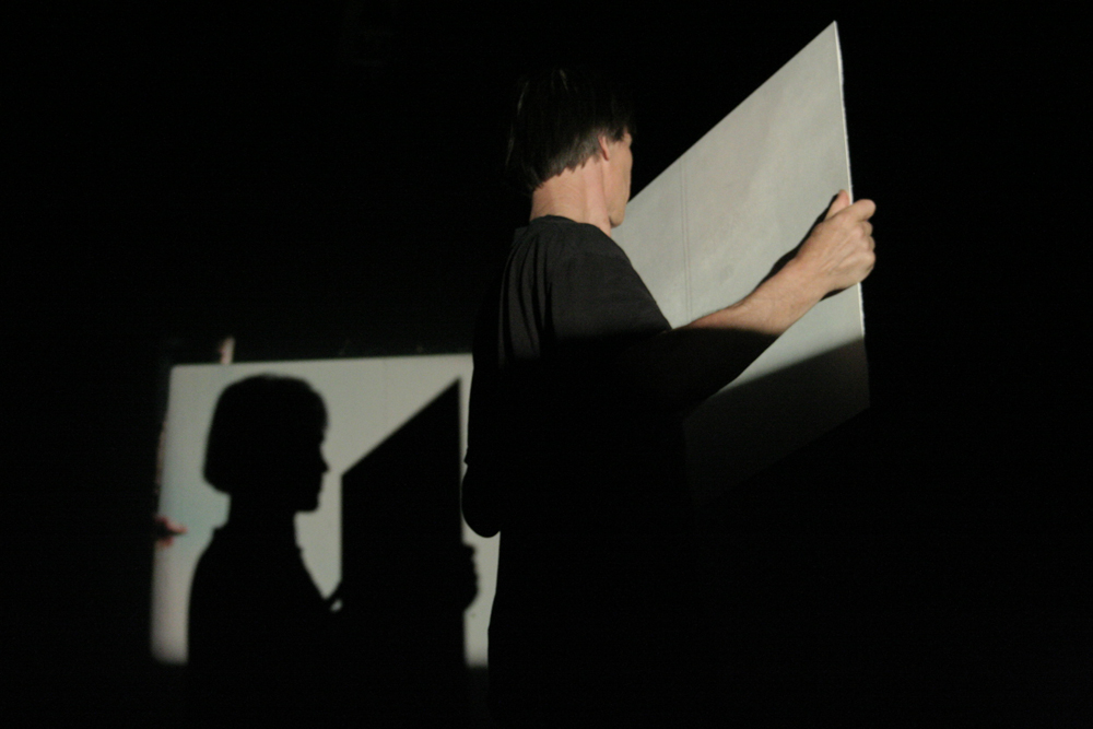 Guy Sherwin holding a mirror, the back of the mirror visible, a shadow