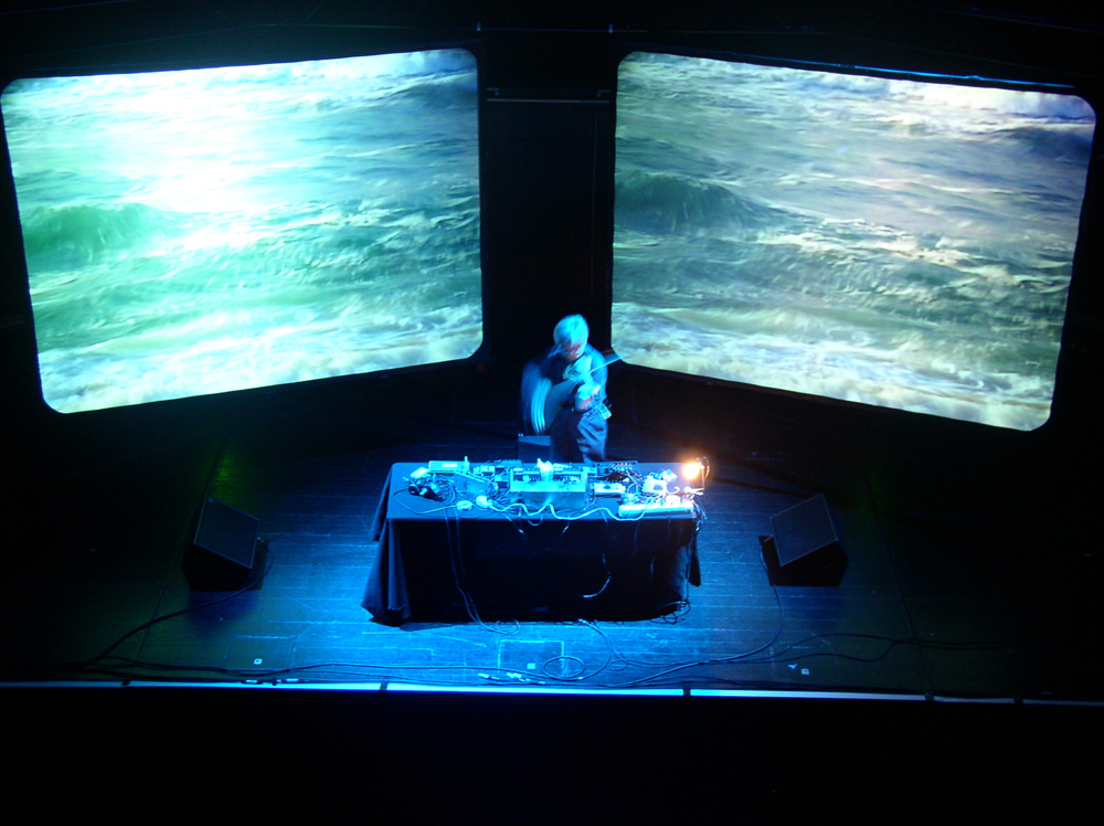 Takehisa Kosugi bowing a violin between two screens showing waves