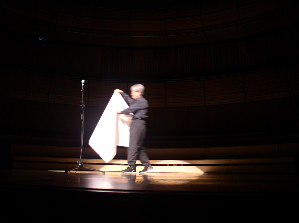 Takehisa Kosugo comes onstage carrying a sheet of paper towards a microphone