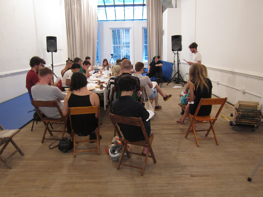 People sit at a long table in a white room as folks listen to a standing figure 