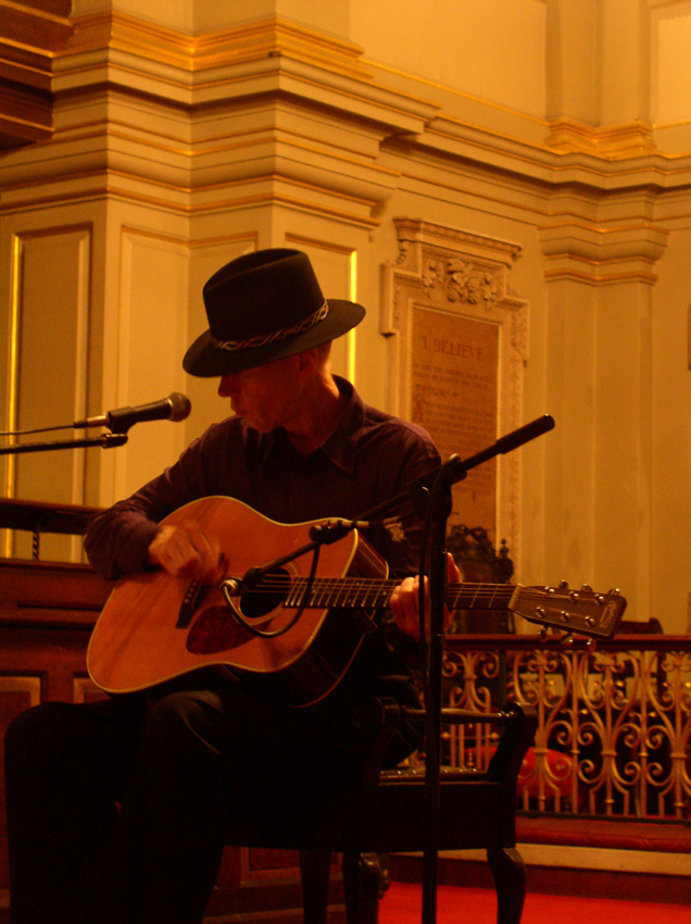 Jandek playing an acoustic guitar and wearing a hat