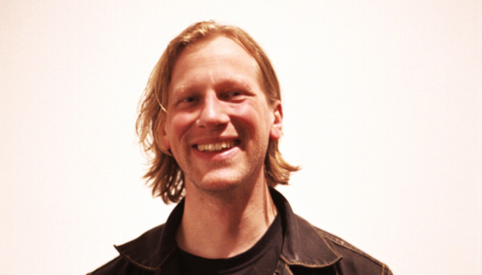 Daniel Menche portrait with black clothes, smiling against a white background 