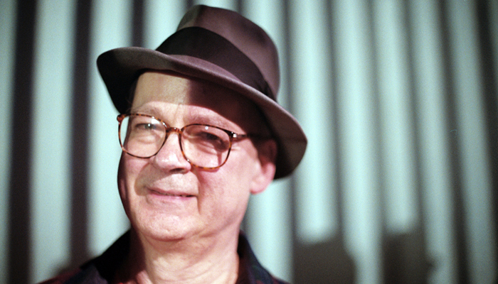 Tony Conrad smiling with glasses and a hat, a little shadow on his face
