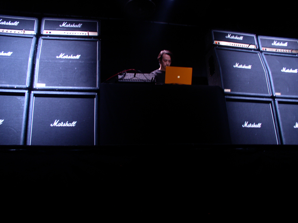 Ryoji Ikeda operating a laptop surrounded by amplifiers