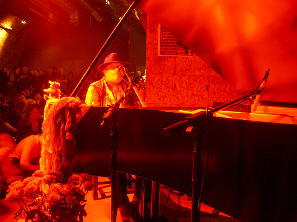 Charlemagne Palestine seated at a piano in red light, small bear to his right