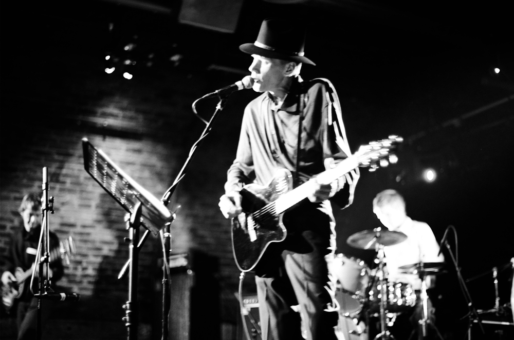 Jandek on stage wearing a hat & playing an electric guitar