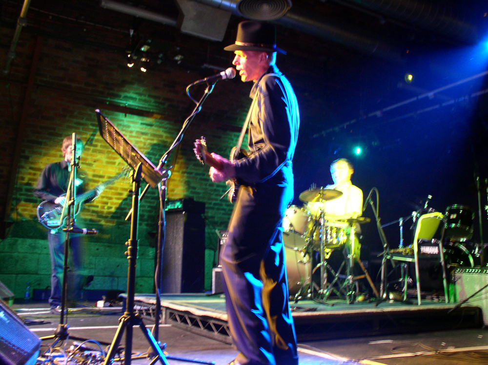 Jandek in a black stetson plays guitar flanked by a drummer and a bassist