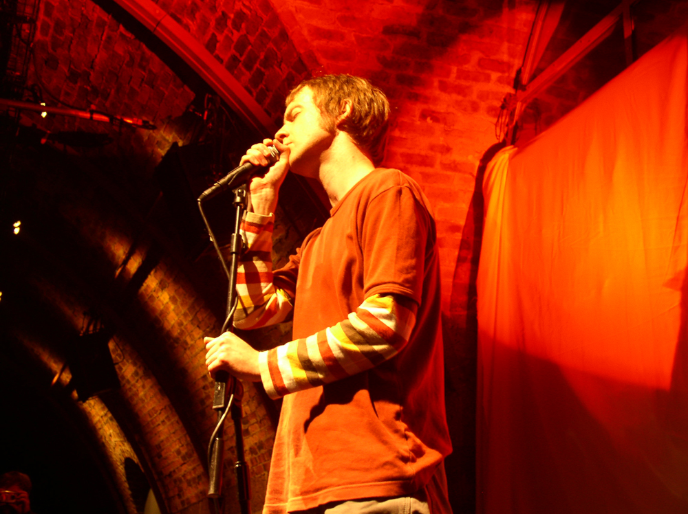 Richard Youngs stands singing into a microphone in orange light