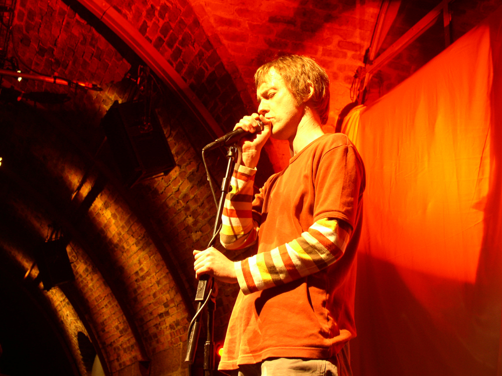 Richard Youngs stands singing into a microphone in orange light