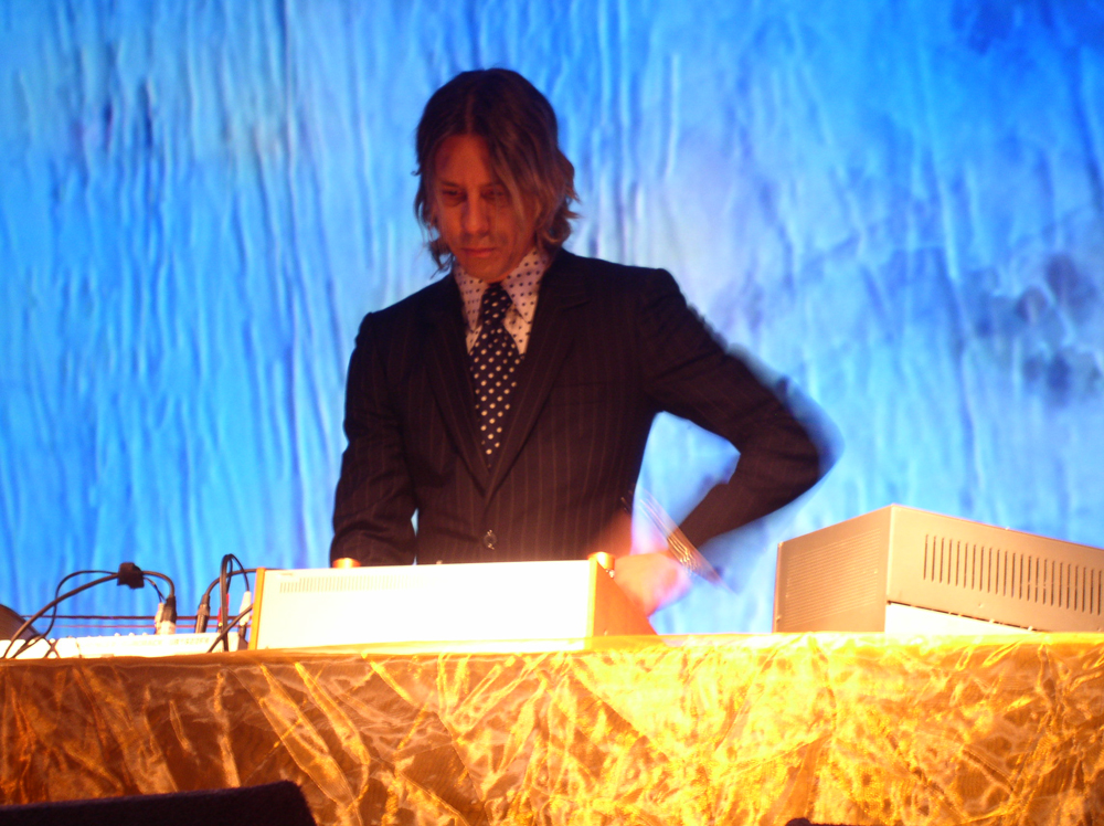 William Basinski in shirt and tie, blue screen behind, gold table to the fore