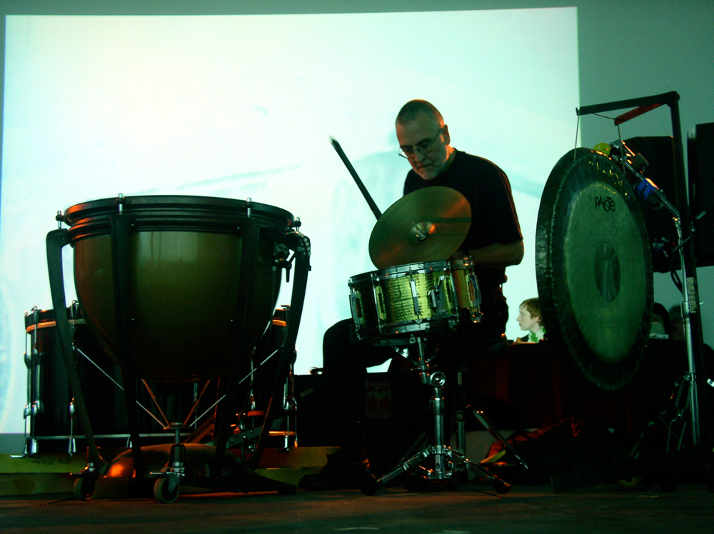Eddie Prevost bowing a cymbal near a snare drum in front of a projection
