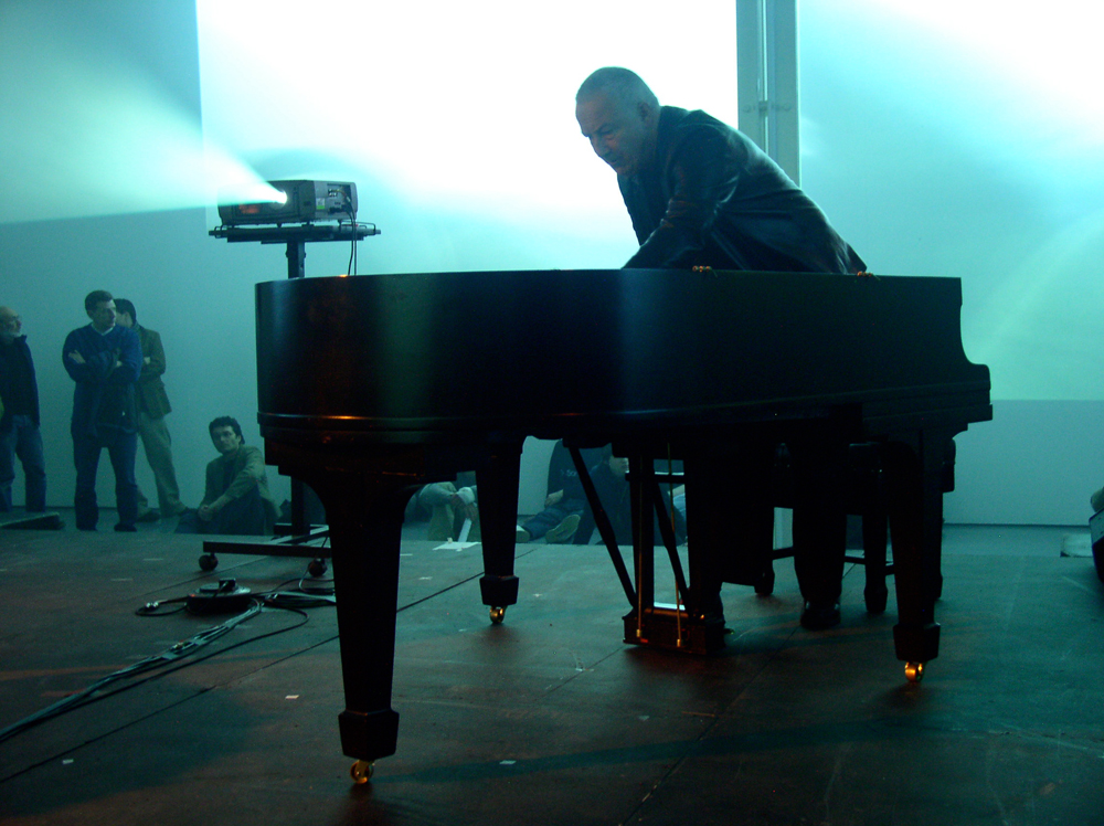 John Tilbury playing a piano in a room full of projections