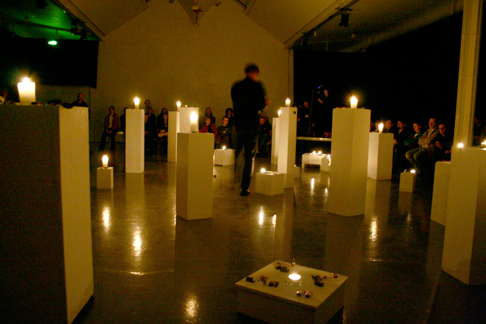 A room full of plinths with candles on top, audience nearby