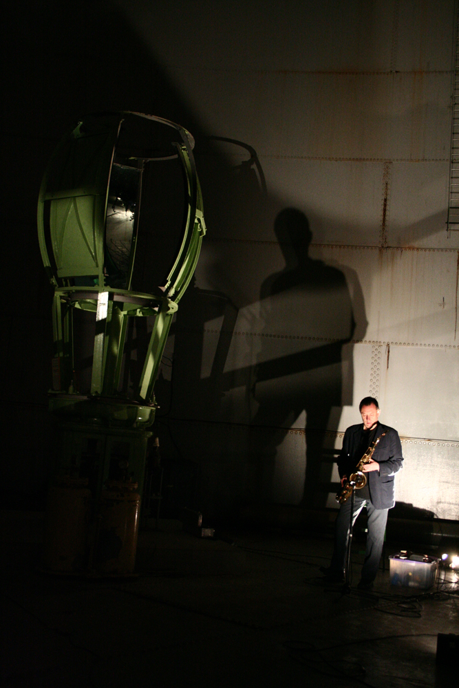 John Butcher playing tenor sax in an oil tank next to old metal machinery