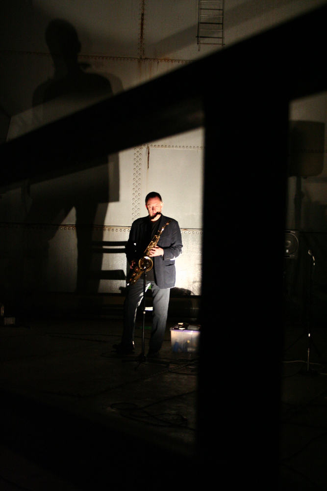 John Butcher holding a tenor saxophone in darkness in an oil tank