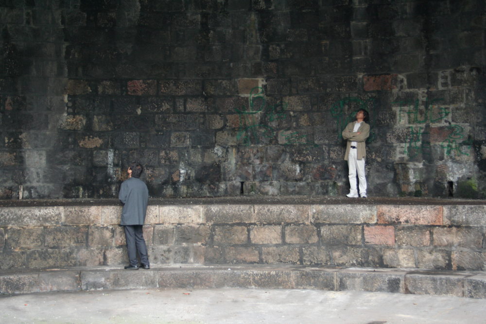 Tamio and Ikuru looking around at a stone walled space with green patches