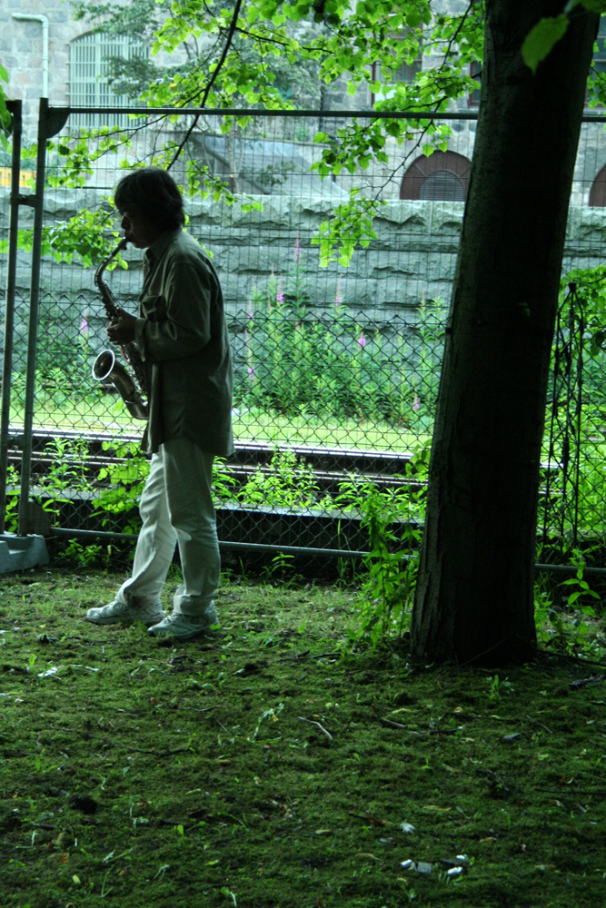 Tamio Shiraishi playing saxophone under a tree in white jeans and coat