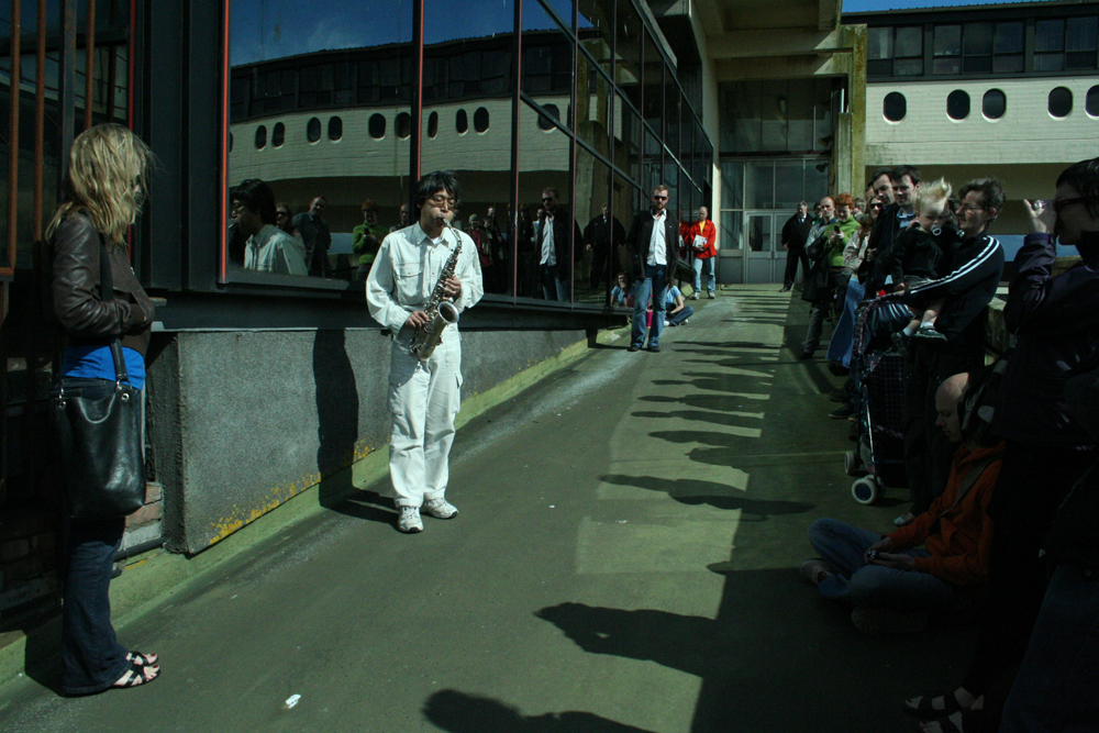 Tamio Shiraishi dressed in white plays saxophone on a walkway in Cumbernauld 