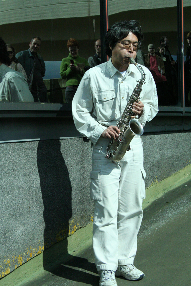 Tamio Shiraishi dressed in white plays saxophone on a walkway in Cumbernauld 