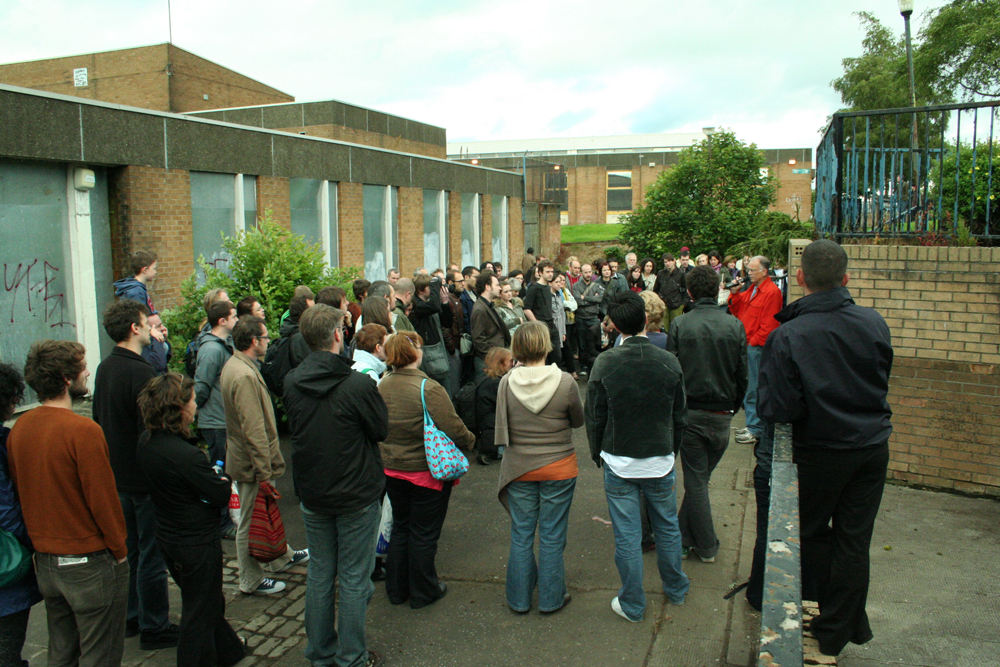 An audience assembled around Denis Wood speaking in Easterhouse