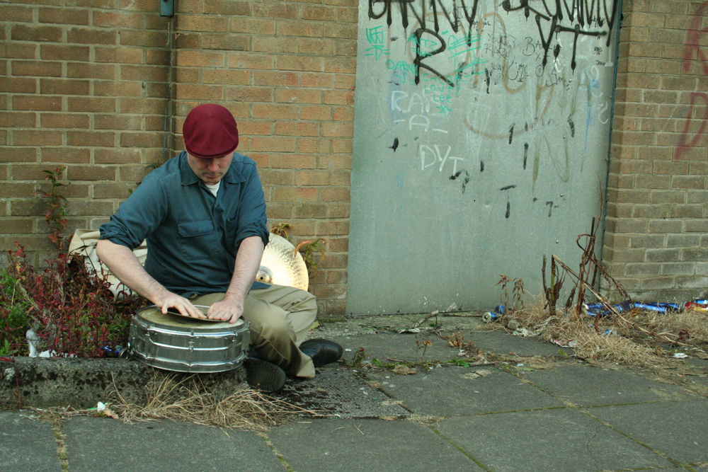 Sean Meehan holding a cymbal against a snare near a metal door, red hat