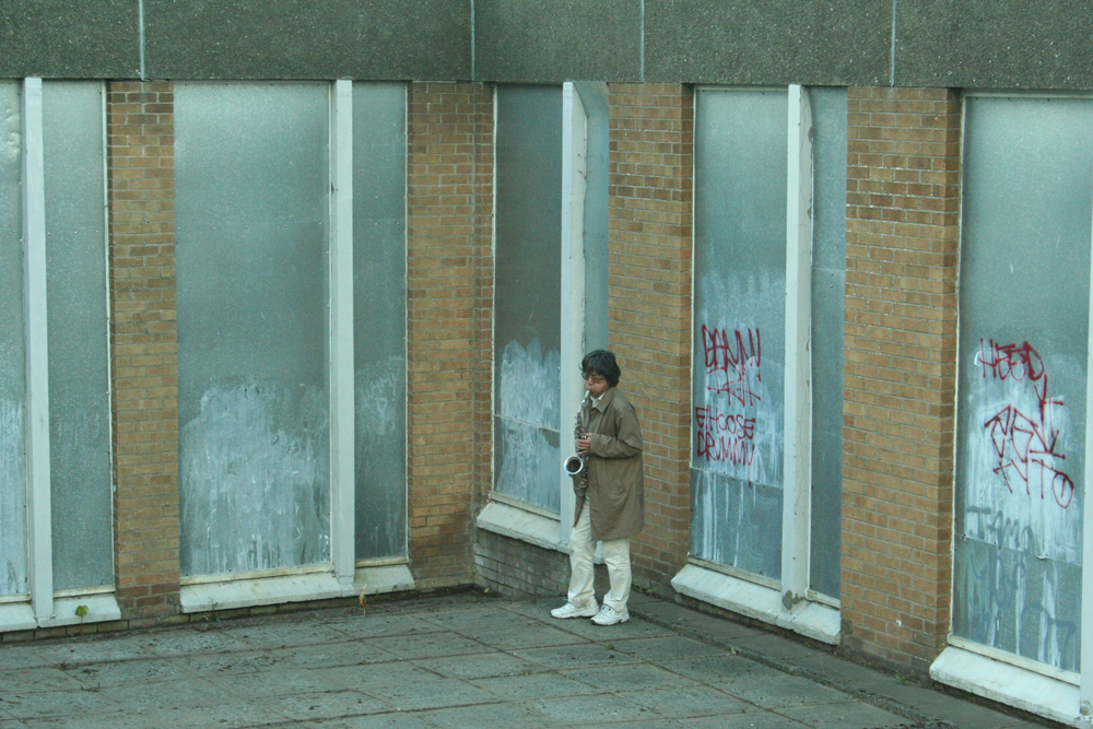 Tamio Shiraishi playing saxophone next to a wall with a series of metal panels