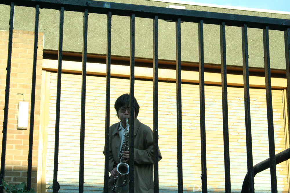 Tamio Shiraishi playing saxophone behind a metal fence