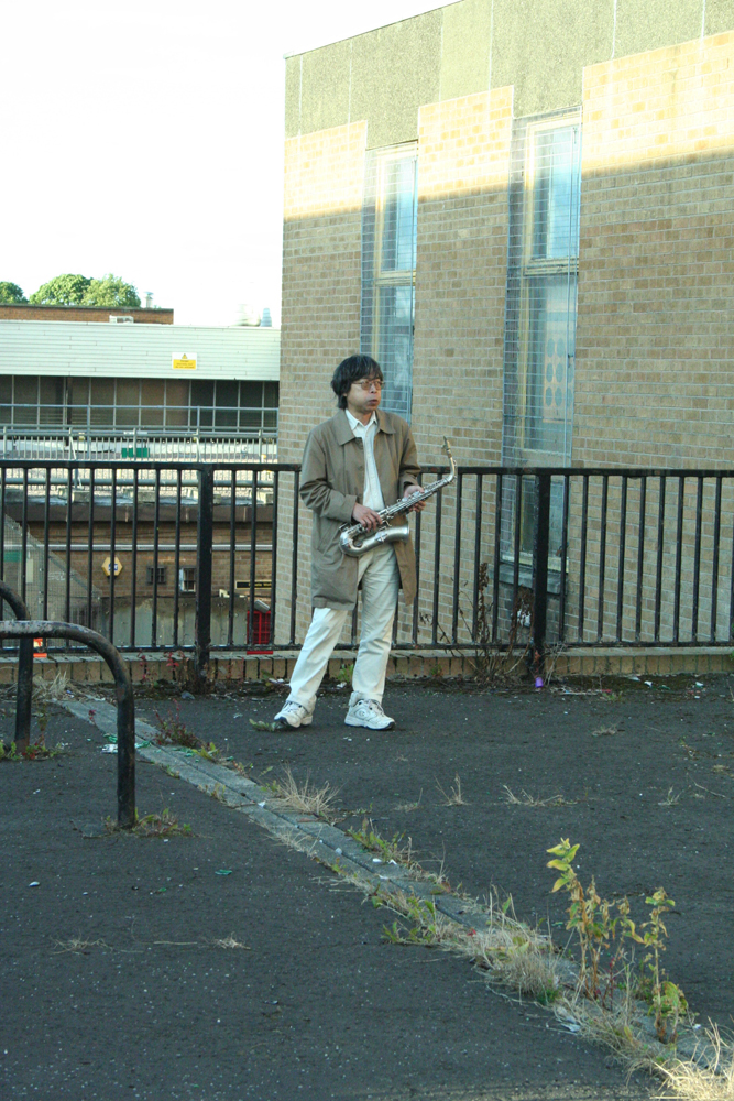 Tamio standing alone near a fence holding a saxophone