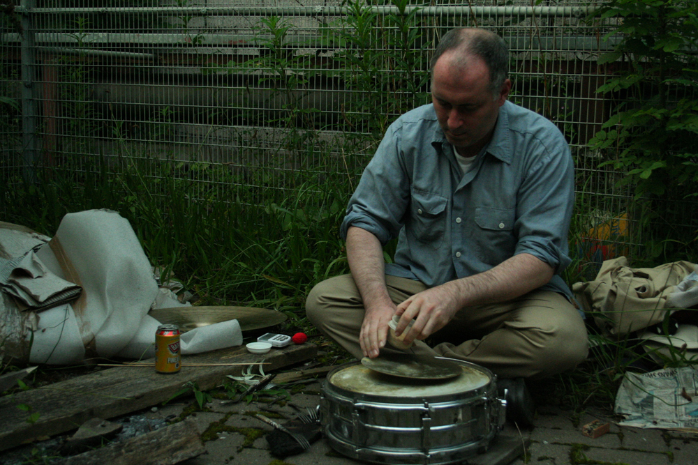 Sean Meehan playing a cymbal on a snare next to a fence