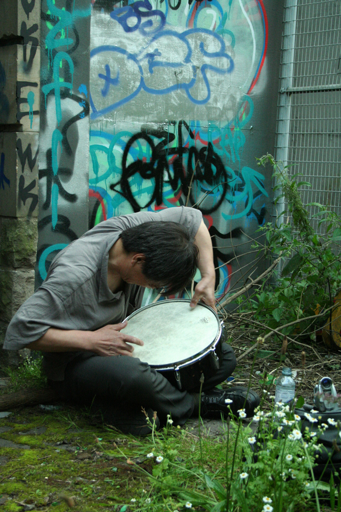 Ikuru Tahehashi playing a snare next to a graffiti covered wall and a fence