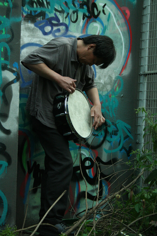 Ikuru Takehashi playing a snare standing against a graffiti covered wall