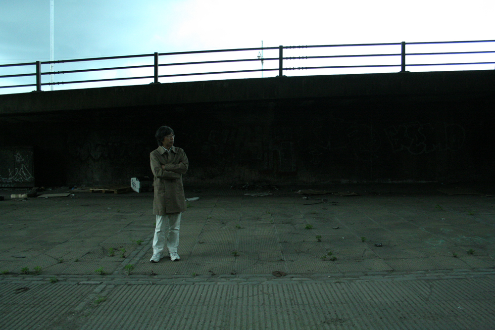 Tamio with folded arms stands by a motorway in a raincoat