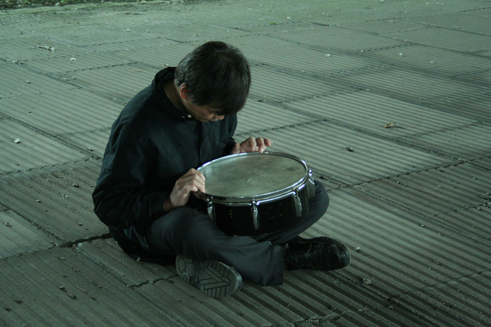 Ikuro Takehashi sitting on ribbed concrete tiles playing a snare