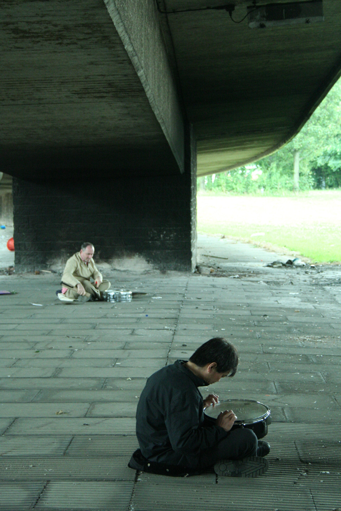 Two men with snare drums playing under a dirty motorway