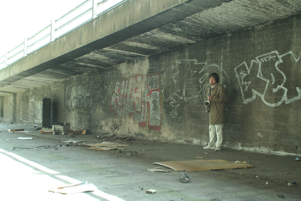 Tamio Shiraishi playing saxophone next to a concrete wall