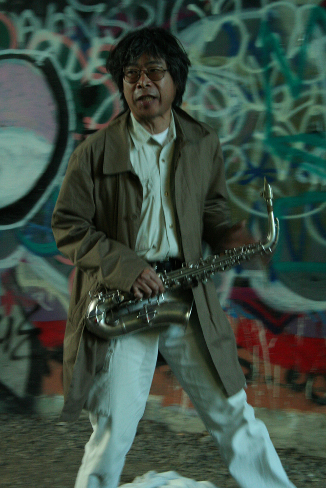Tamio Shiraishi with saxophone against graffiti covered walls, white jeans