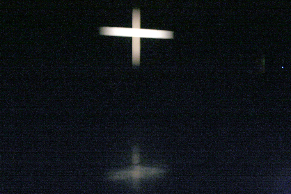 A white cross projected on a screen is reflected on a shiny floor below